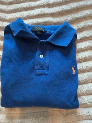 Blå pikétröja från Ralph Lauren - Klassisk blå pikétröja från Ralph Lauren med orange broderad logga på bröstet. Bra skick använt fåtal gånger