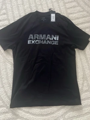 Svart Armani Exchange t-shirt - Snygg svart t-shirt från Armani Exchange med stort logotryck i vitt på bröstet. Modellen har rund hals och korta ärmar. Slim fit för en modern look. Perfekt till jeans eller shorts för en clean och stilren outfit.