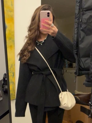 Svart omlott kappa med knyt band  - Stilren svart kappa i omlottmodell med bälte i midjan. Jackan har lång ärm och en rak, loose passform som ger en avslappnad men chic look. Perfekt att slänga över en outfit för en trendig vibe.