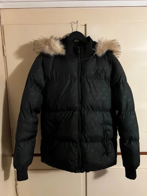 Svart pufferjacka från SikSilk - Svart pufferjacka från SikSilk med avtagbar huva och fuskpälsdetalj. Jackan har diskret logga på bröstet, quiltad design och dragkedja framtill. Perfekt för kalla dagar med sin tjocka vaddering och långa ärmar.
