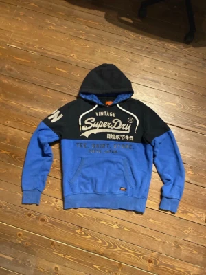 Superdry hoodie -  Superdry Japan hoodie i blått och svart med stor logga och text på bröstet samt japanska tecken. Huva med mönstrat innerfoder. Fint skick