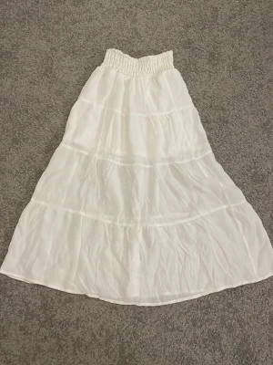Vit långkjol från H&M med volanger - Säljer en vit långkjol från H&M med bred resår i midjan och flera volanglager. Kjolen är luftig och har en lätt, flowy känsla som passar perfekt till varma dagar. Enkel att matcha med olika toppar för en chill och stilren look.