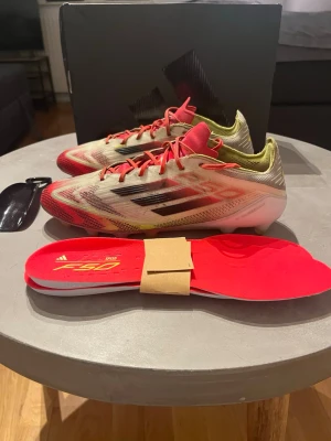 Adidas F50 fotbollsskor röd/vit - Säljer ett par Adidas F50 fotbollsskor i rött, vitt och svart med gula detaljer. Skorna har snörning, platt sula med dobbar och är tillverkade i syntetmaterial. Perfekta för dig som vill ha lätta och snabba skor på planen. Innersulor och skohorn medföljer.