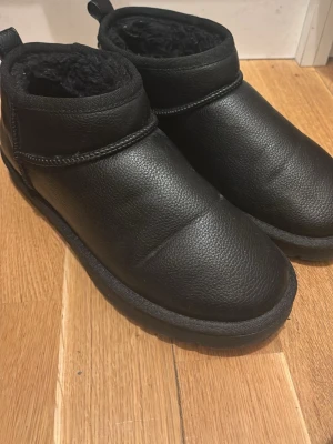 Svarta tofflor i skinnimitation - Säljer ett par svarta tofflor/ uggs med rund tå och platt sula. Utsidan är i skinnimitation och insidan är fodrad med mjuk syntetpäls för extra värme. Perfekta för kalla dagar och har en enkel, stilren design.