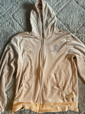 Juice Couture peach hoodie - Säljer en peach färgad hoodie från Juicy Couture med glittrande JC-logga i strass på bröstet. Hoodien har dragkedja, kängurufickor och en stor huva. Perfekt för dig som gillar en mjuk och lyxig vibe.
