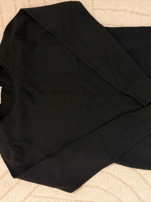 Svart tröja med struktur från Flora - Snygg svart långärmad tröja från Flora med diskret prickig struktur i tyget och en liten bröstficka. Tröjan har rund halsringning och ribbade muddar vid ärmslut. Perfekt till jeans eller kjol för en clean look.