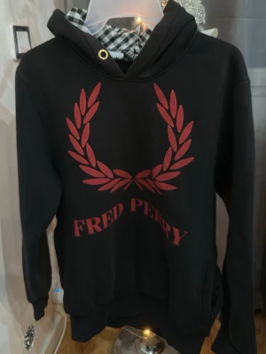 Svart Fred Perry hoodie med röd logga - Svart hoodie från Fred Perry med stor röd lagerkrans och text på bröstet. Klassisk huva med snörning och ribbade muddar. Perfekt för en avslappnad och stilren look.
