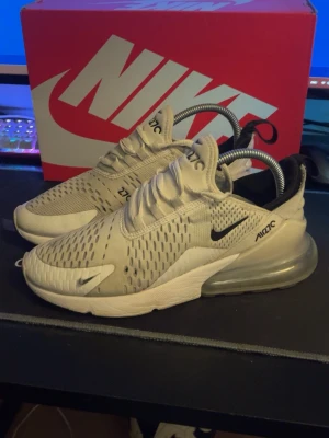 Nike Air Max 270 vita sneakers - Nike Air Max 270 sneakers i vitt med svarta detaljer och stor synlig Air Max-sula i hälen. Ovandelen är i mesh för bra andningsförmåga och har en sportig, modern design med tydlig Nike-logga på sidan. Perfekta för en streetwear-look.