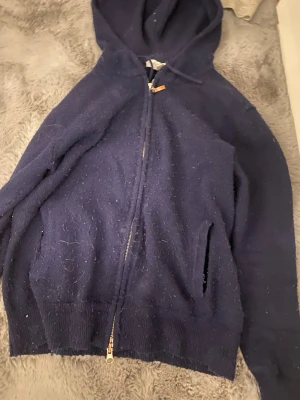 Mörkblå hoodie från Ahlquist Clothing - Mysig mörkblå hoodie från Ahlquist Clothing med dragkedja framtill och två fickor. Tillverkad i ett mjukt, stickat material som känns skönt mot huden. Klassisk huva med dragsko och ribbade muddar vid ärmslut och nederkant. Perfekt för en avslappnad look.