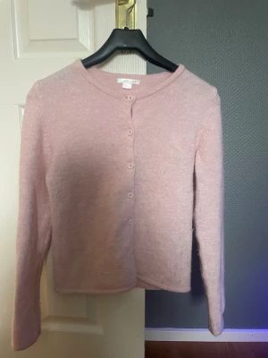 Ljusrosa kofta från Gina Tricot - Mysig ljusrosa kofta från Gina Tricot med rund halsringning och knappar framtill. Koftan har lång ärm och är gjord i ett mjukt, fluffigt material som känns skönt mot huden. Perfekt att slänga över en topp för en söt och enkel look.