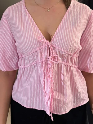 Rosa randig blus med knyt framtill - Supersöt rosa och vit randig blus med v-ringning och knyt framtill. Blusen har korta, lite vidare ärmar och ett luftigt, lätt material som känns perfekt för varmare dagar. Passformen är normal och toppen har en avslappnad, feminin look.