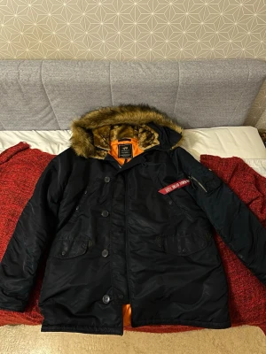 Alpha industries  - Svart parkasjacka från Alpha Industries med avtagbar fuskpäls runt huvan och orange innerfoder. Jackan har flera fickor, dragkedja och knappar framtill samt en ikonisk röd tag på ärmen. Perfekt för kalla dagar och riktigt snygg streetstil.
