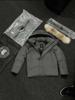 Grå dunjacka från Canada Goose - Snygg grå dunjacka från Canada Goose med avtagbar pälskant till huvan. Jackan har en quiltad design, långa ärmar och dragkedja med knappar framtill. Perfekt för kalla vinterdagar och riktigt varm. Klassisk logga på ärmen.