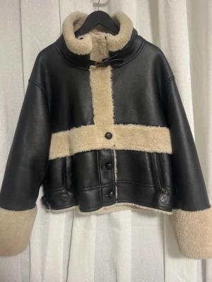 Svart skinnjacka med beige päls - Snygg svart skinnjacka från Vegancode med beige fuskpälsfoder och detaljer. Jackan har bred krage, knappar och dragkedja samt croppad passform. Perfekt för dig som vill ha en trendig look med kontrasterande färger och mysig känsla.