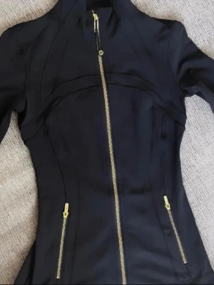 Svart Define jacka från Lululemon - Snygg svart vindjacka från Lululemon med figurnära passform. Jackan har guldfärgade dragkedjor framtill och på fickorna, samt hög krage. Tillverkad i ett stretchigt syntetmaterial som passar perfekt till träning eller vardagsoutfits. Size 2 