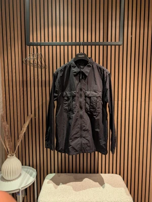 Svart overshirt från H&M - Svart overshirt från H&M med dragkedja framtill och två stora bröstfickor. Jackan har klassisk krage och är långärmad. Materialet känns lätt och passar perfekt för lager-på-lager-stil.