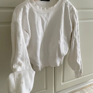 Vit stickad tröja med pärldetaljer - Trendig vit stickad tröja från Chiquelle med rund halsringning och ribbade muddar. Långa ärmar med dekorativa pärlor längs axlar och ärmar ger en unik touch. Perfekt för dig som vill ha en stilren men ändå detaljerad look.