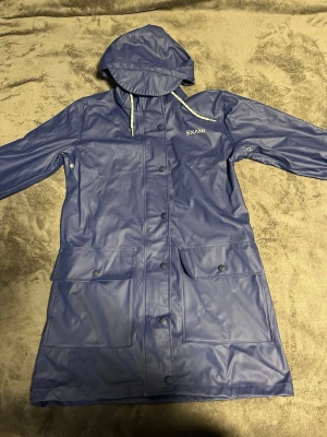 Blå regnjacka med huva från Exani - Snygg blå regnjacka från Exani med huva och dragsko. Jackan har långa ärmar, tryckknappar framtill och två stora fickor med lock. Tillverkad i vattenavvisande syntetmaterial, perfekt för regniga dagar.