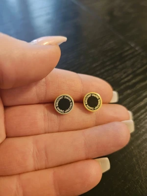 Marc Jacobs svarta guldpläterade örhängen - Säljer ett par runda örhängen från Marc Jacobs i svart och guld. Örhängena har en klassisk design med svart emalj i mitten och guldfärgad kant där märkesnamnet står runt om. Perfekta för dig som gillar stilrena accessoarer med logga.
