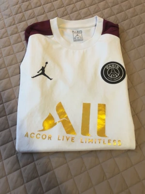 PSG x Jordan vit matchtröja - Säljer en vit PSG matchtröja i samarbete med Jordan. Tröjan har vinröda axlar, svart Paris Saint-Germain-märke på bröstet och guldfärgad 'Accor Live Limitless'-logga framtill. Tillverkad i lätt och bekvämt material, perfekt för fotboll eller träning.