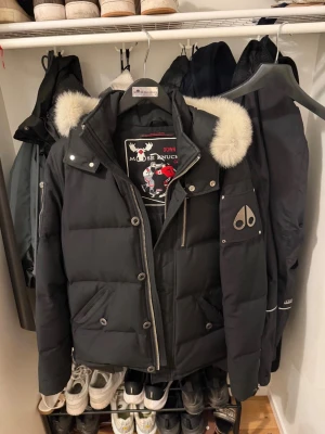 Svart dunjacka från Moose Knuckles - Snygg svart dunjacka från Moose Knuckles med puffer-design och avtagbar huva med fluffig vit päls. Jackan har dragkedja, knappar och flera fickor framtill samt logga på ärmen. Perfekt för kalla vinterdagar och riktigt varm.finns i stockholm 