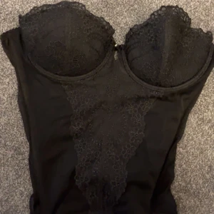 Svart spetskorsett med bygel - Säljer en svart korsett med bygel och vacker spetsdetalj över kuporna och framtill. Toppen har hakknäppning bak och en figurnära passform. Storlek:75b 