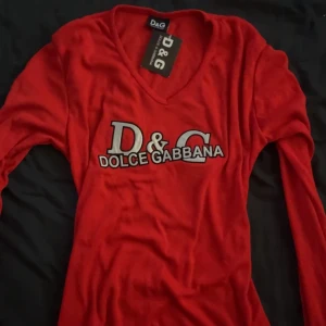 Röd D&G långärmad tröja med glitter tryck - Snygg röd långärmad tröja från Dolce & Gabbana med v-ringning och stort glittrigt D&G-tryck framtill. Dm om du har funderingar 💘
