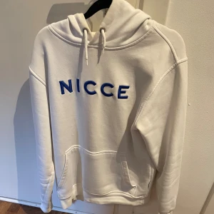 Vit hoodie från NICCE med blå text - Snygg vit hoodie från NICCE med blå broderad logga på bröstet. Tröjan har huva med snörning och en stor magficka. Perfekt för dig som gillar en clean och stilren look. Mjuk och skön, passar till allt.