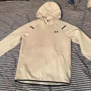 Vit hoodie från Under Armour M - Snygg vit hoodie från Under Armour med svart logga på bröstet och svarta detaljer vid axlarna. Tröjan har huva, lång ärm och en stor magficka. Perfekt för chill eller träning, riktigt clean look och skön passform.