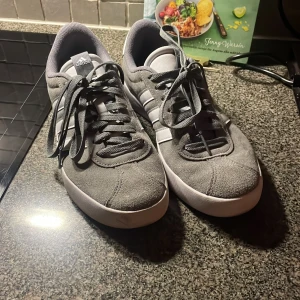 Gråa Adidas sneakers i mocka - Snygga grå sneakers från Adidas med klassiska vita ränder på sidorna och vit sula. Ovandelen är i mjuk mocka och skorna har rund tå och snörning. Perfekta för en clean och sportig look.