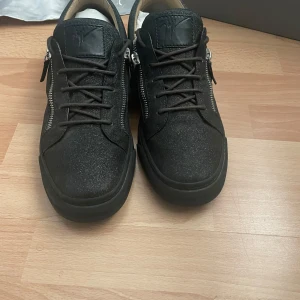 Svarta sneakers från Giuseppe Zanotti - Snygga svarta sneakers från Giuseppe Zanotti med glittrig finish, snörning och silverfärgade dragkedjor på sidorna. Skorna har rund tå, platt sula och är tillverkade i mocka med detaljer i skinn. Perfekta för dig som gillar exklusiv streetstyle.