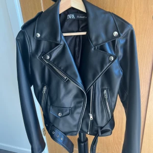 Svart skinnjacka från Zara, M - Snygg svart skinnjacka från Zara i klassisk bikerstil med silvriga dragkedjor, knappar och bälte i midjan. Jackan har långa ärmar, bred krage och flera fickor med dragkedja. Perfekt för dig som vill ha en edgy och stilren look.