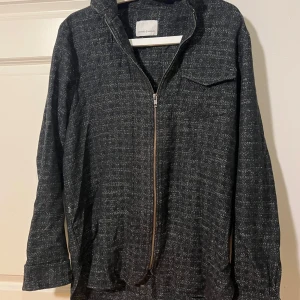 Svart overshirt från Samsøe Samsøe - Stilren svart overshirt från Samsøe Samsøe med diskret rutigt mönster och hel dragkedja framtill. Jackan har en bröstficka, klassisk krage och långa ärmar. Perfekt lager-på-lager-plagg med en lite loose passform.