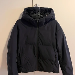 Svart pufferjacka från Uniqlo  - Svart pufferjacka från Uniqlo i storlek XS. Jackan har huva, dragkedja framtill och är långärmad. Materialet är syntet och jackan är både vind- och vattenavvisande. Passformen är loose och modellen är lite croppad. Perfekt för kalla dagar.