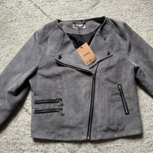 Grå mocka bikerjacka från H&M - Snygg grå bikerjacka i mockaimitation som från början är köpt på H&M men sedan har jag köpt den på sellpy. Jackan har flera dragkedjor framtill, snedställd dragkedja och coola detaljer. Perfekt för dig som vill ha en stilren men ändå tuff look.