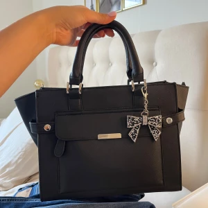 Svart handväska med rosett-detalj - Snygg svart handväska som jag aldrig har använt , snygg till skola och annat . Mitt är 28b,24H och 12D