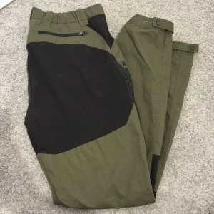 Gröna och svarta cargopants med dragkedjor - Snygga cargopants i olivgrönt och svart med flera dragkedjefickor och förstärkta partier. Byxorna har elastisk midja, bälteshällor och tryckknappar vid benslut. 