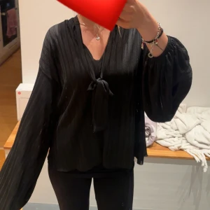 Svart blus med knyt och vida ärmar - Säljer en svart blus med snygg knytning framtill och v-ringning. Blusen har vida, långa ärmar och ett lätt glansigt tyg med diskreta ränder. Perfekt för dig som gillar en stilren men ändå trendig look.