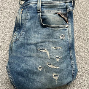 Replay jeans - Snygga blå jeans med slitningar för en trendig look från replay. De har en klassisk femficksdesign och en brun läderdetalj vid midjan. Perfekta för en avslappnad stil. Enda grejen är att lappen är lite sönder i sömmen