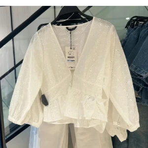 Vit blus med broderade detaljer - Superfin slutsåld vit blus från Zara med broderade blommor och v-ringning. Blusen har volangkant nertill, ballongärmar och knappar i midjan. Superbra skick då den knappt är använd. 