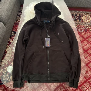 Svart hoodie från Polo Ralph Lauren - Svart hoodie från Polo Ralph Lauren med klassisk broderad logga på bröstet. Tröjan har huva med snörning, dragkedja framtill och två fickor. Tillverkad i mjuk bomull med ribbade muddar vid ärmslut och nederkant. Perfekt för chill dagar och streetwear.