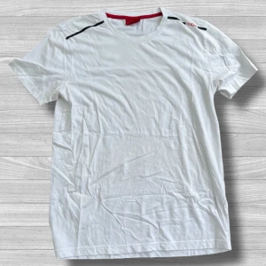 Hugo Boss - Vit t-shirt från Hugo Boss med rund hals och diskreta svarta detaljer på axlarna. Klassisk passform och mjukt bomullsmaterial som känns skönt mot huden. Röd etikett i nacken ger en snygg kontrast. Perfekt för en clean och stilren look.