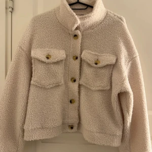 Beige teddyjacka med stora fickor - Supermjuk och fluffig beige teddyjacka med stora bröstfickor och knappar framtill. Jackan har en boxig passform och hög krage, perfekt för lager-på-lager. Snygg att styla till både jeans och kjol för en chill look.