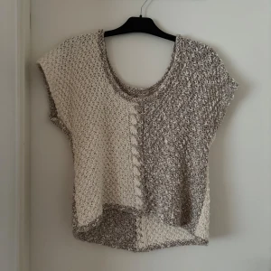 Beige och brun stickad topp - Unik stickad linnetopp i beige och grått med grov struktur och snygg kabeldetalj framtill. Toppen är ärmlös och har en avslappnad passform med rund halsringning. Perfekt för lager-på-lager eller som statement-piece till jeans.
