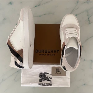 Burberry vita sneakers med rutig detalj - Snygga vita sneakers från Burberry med klassiskt rutig panel i beige, svart och brunt på sidorna. Skorna har ovandel i skinn och mocka, vita snören och platt sula. Perfekt för dig som vill ha en stilren men ändå ikonisk look.