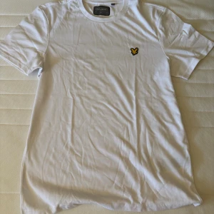 Vit t-shirt från Lyle & Scott - Vit t-shirt från Lyle & Scott med klassisk rund hals och korta ärmar. På bröstet sitter den ikoniska gula fågel-loggan. T-shirten är gjord i mjuk bomull och har en clean, stilren look som passar perfekt till jeans eller shorts.