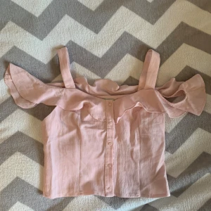 Ljusrosa offshoulder blus med volang - Supersöt ljusrosa blus med offshoulder och breda axelband. Toppen har volangdetalj längs överdelen och knappar framtill. Perfekt för en somrig look och lätt att matcha med jeans eller kjol.