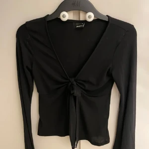 Svart omlottopp med knyt från Gina Tricot - Svart långärmad omlottopp från Gina Tricot med djup v-ringning och knyt framtill. Toppens passform är slim och den är tillverkad i mjuk bomullsmix. Perfekt för dig som gillar stilrena och trendiga plagg.