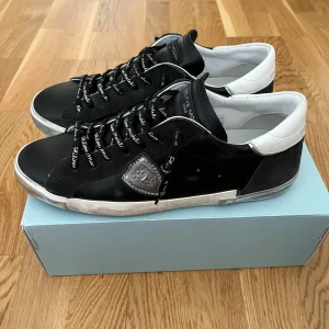 Philippe Model svarta sneakers - Svarta sneakers från Philippe Model med vita detaljer och logga på sidan. Skorna har snörning med text på skosnörena och en vit hälkappa. Ovandelen är i skinn och mocka, med en platt sula och rund tå. Snygg och urban stil som passar till många outfits.