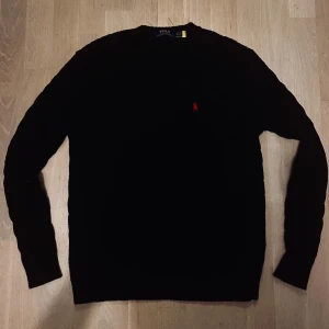  Ralph Lauren knit - Svart kabelstickad tröja från Polo Ralph Lauren med klassisk röd broderad logga på bröstet. Tröjan har lång ärm och rund halsringning, perfekt för dig som gillar stilren och tidlös design.